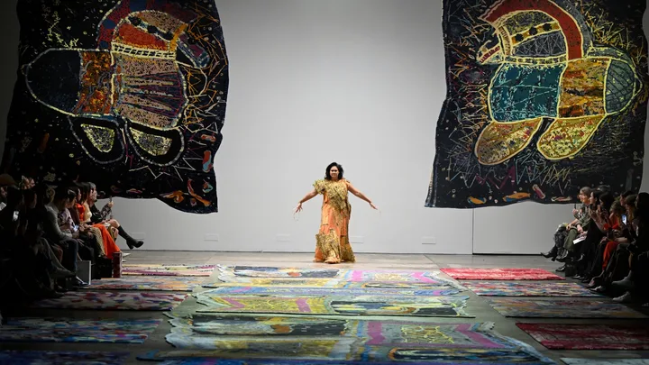 Model walks runway with vibrant abstract art; audience sits on either side.