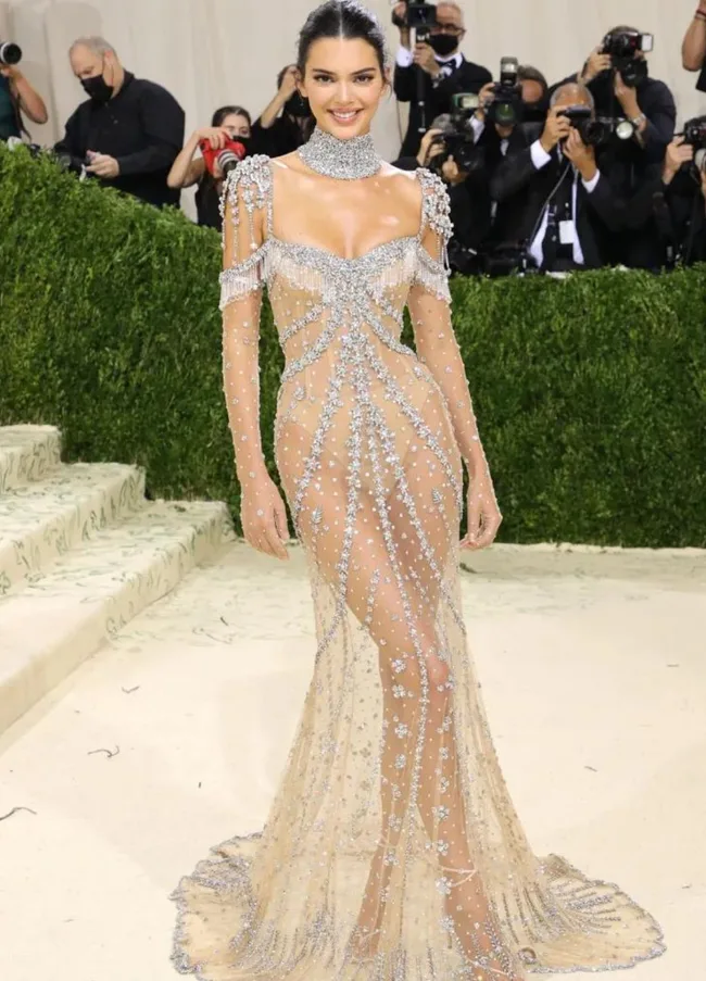 Person in a sheer, embellished gown poses on a beige carpet with photographers in the background.