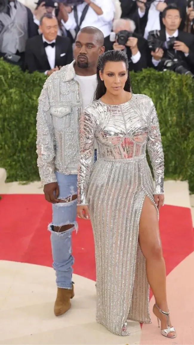 Two individuals in stylish silver outfits pose on a red carpet, surrounded by photographers at an event.