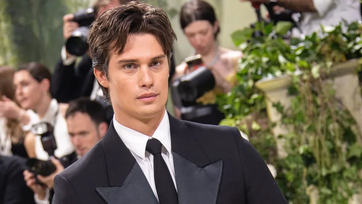 A person in a tuxedo at a formal event with photographers in the background.