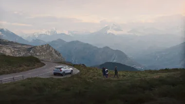 A silver car parked on a winding mountain road with three people walking nearby, snow-capped peaks in the background.