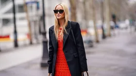 Woman in sunglasses wearing a black jacket over a red dress, walking down a city street.