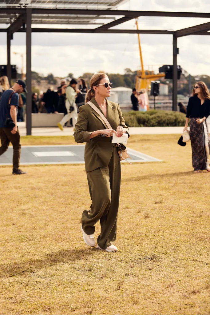 a woman with a blonde ponytail is photographed walking across some grass. She wears a stylish olive suit with sneakers