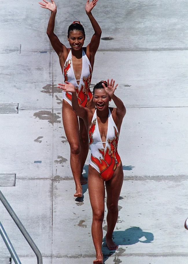 Two synchronized swimmers in matching flame-patterned swimsuits waving and smiling on a poolside deck.