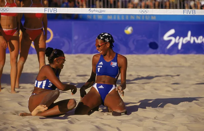 Two beach volleyball players in blue Cuban uniforms smiling on sand at the 2000 Sydney Olympics.