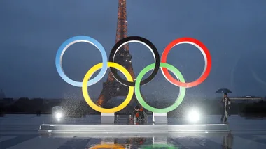 a shot of the olympic rings, lit up at night, with the Eiffel Tower as a shadow in the background, taken ahead of the 2024 Olympics in Paris