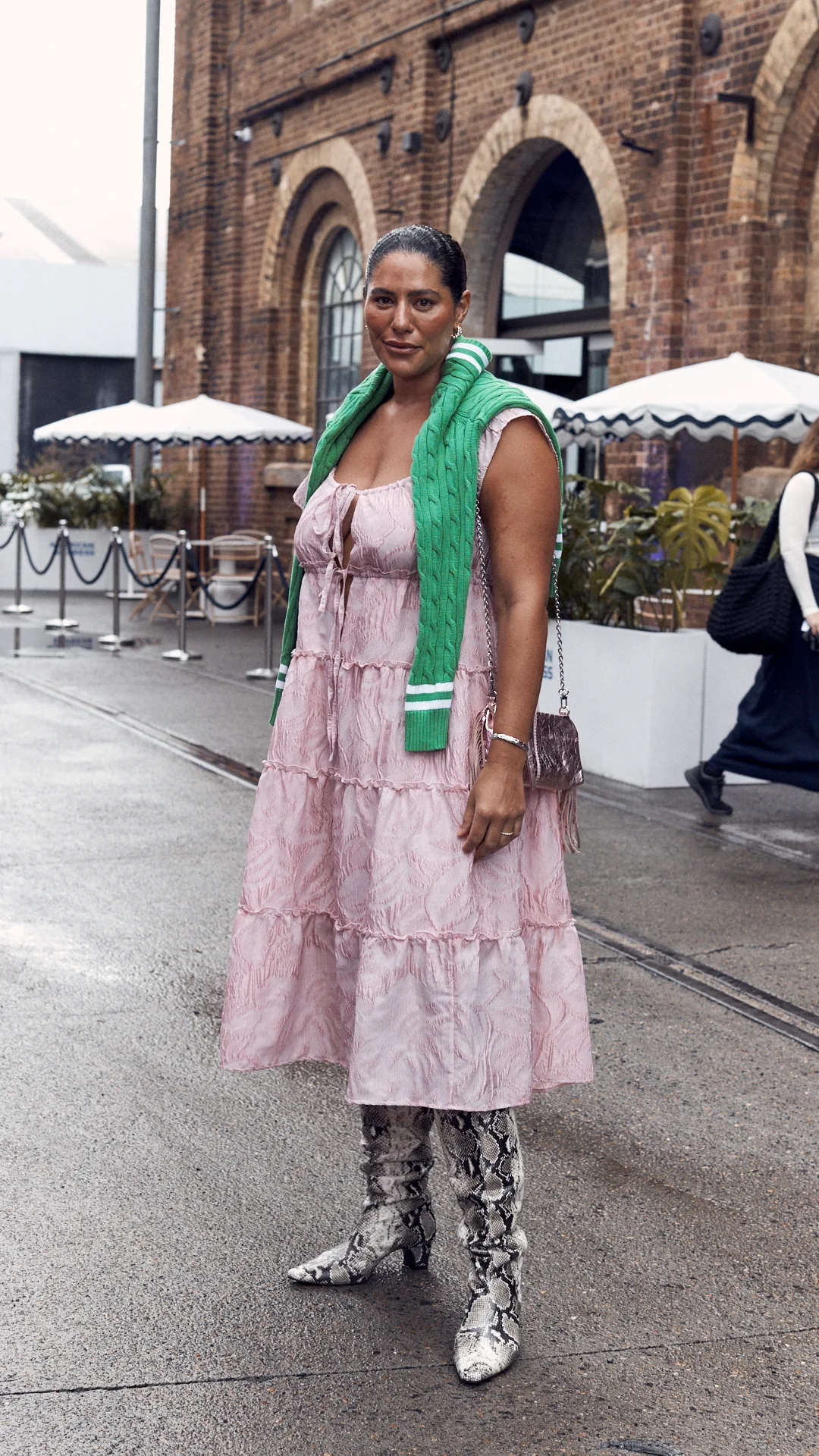 aa stylish woman wears her brown hair slicked back into a ponytail. She faces the camera, wearing a pink calf-length dress and knee-high boots, as well as a green knit jumper thrown around her shoulders