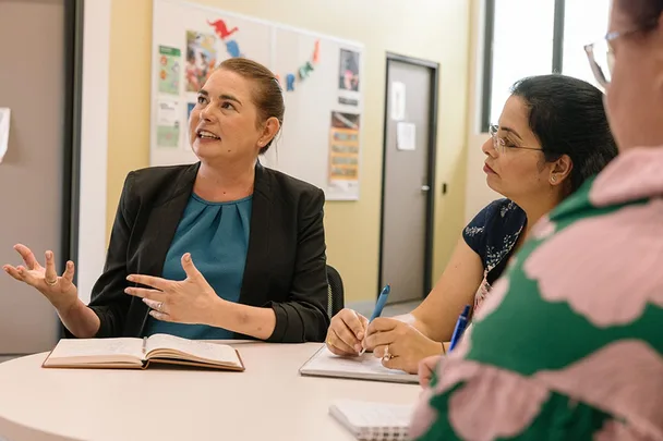 Three women discussing around a table, one speaking passionately with a notebook open.