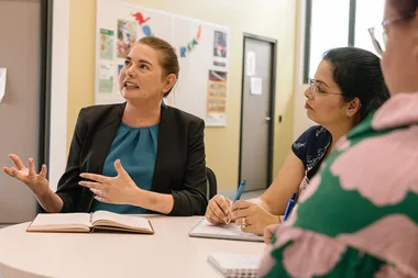 Three women discussing around a table, one speaking passionately with a notebook open.