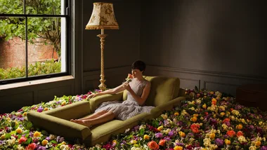 Woman in a dress sits on a green sofa surrounded by colorful flowers, holding a rose, next to a window and lamp.