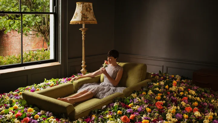 Woman in a dress sits on a green sofa surrounded by colorful flowers, holding a rose, next to a window and lamp.
