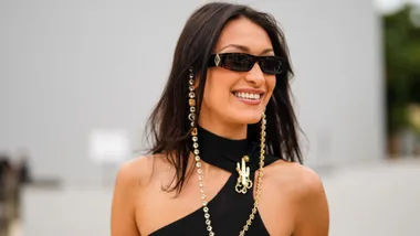 A woman in sunglasses smiles, wearing a black top with gold and crystal chain accessories outdoors.