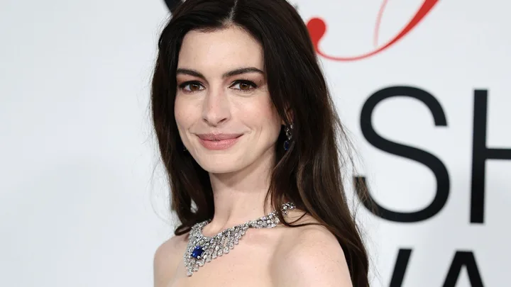 A close up of anne hathaway at the 2023 cfda awards. She looks toward the camera,smiling slightly with her brown hair loose around her shoulders and a large diamond necklace on