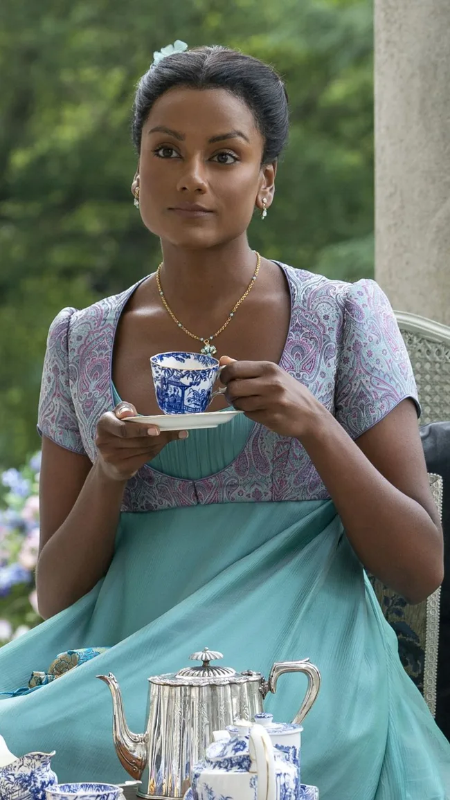 A woman in Regency attire sips tea from a blue and white cup, with a matching teapot on the table, set outdoors.