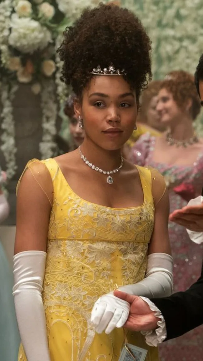 Young woman in yellow gown and tiara at a formal event, wearing a diamond necklace and white gloves, holding an arm.