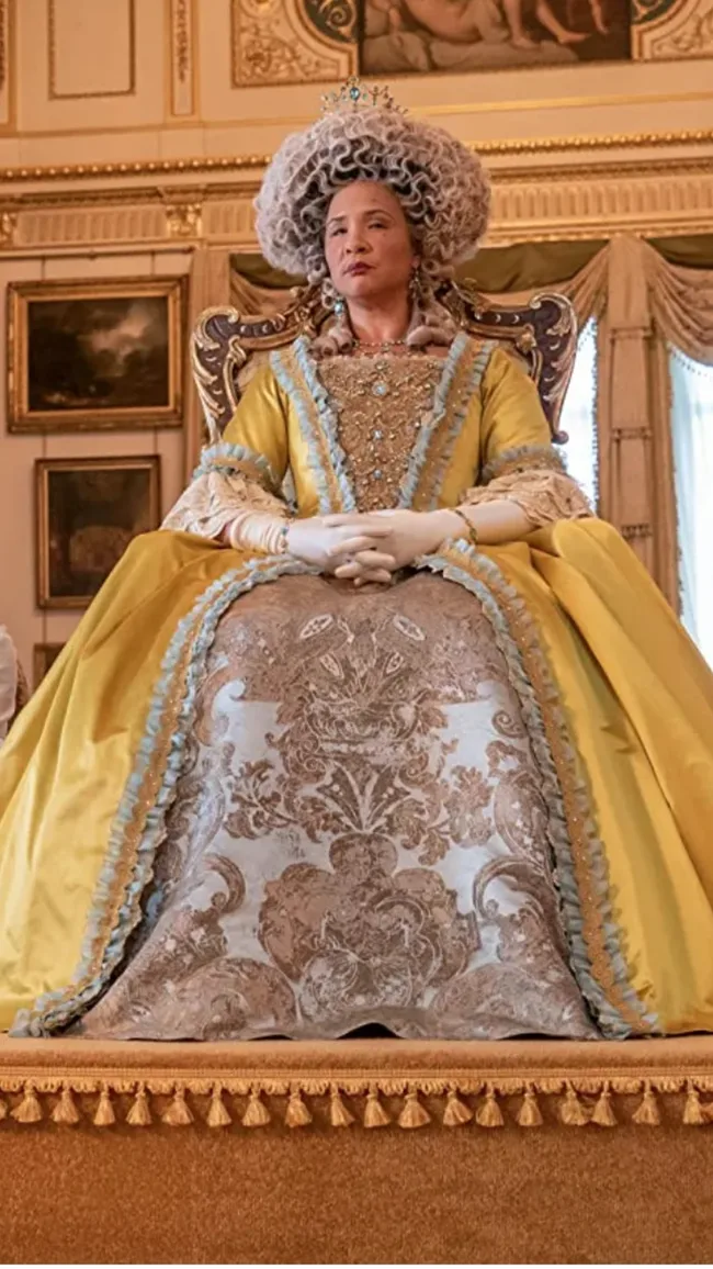 A regal character in an ornate yellow dress and elaborate hairstyle, seated on a throne in an opulent room.