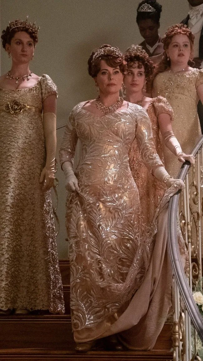 Elegant women in ornate period dresses descending a staircase, wearing tiaras and jewelry.