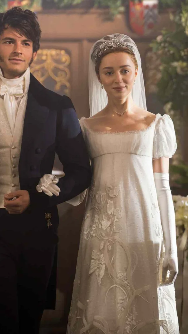 Phoebe Dynevor as Daphne Bridgerton. She wears a wedding dress and holds the arm of Anthony Bridgerton, played by Jonathan Bailey