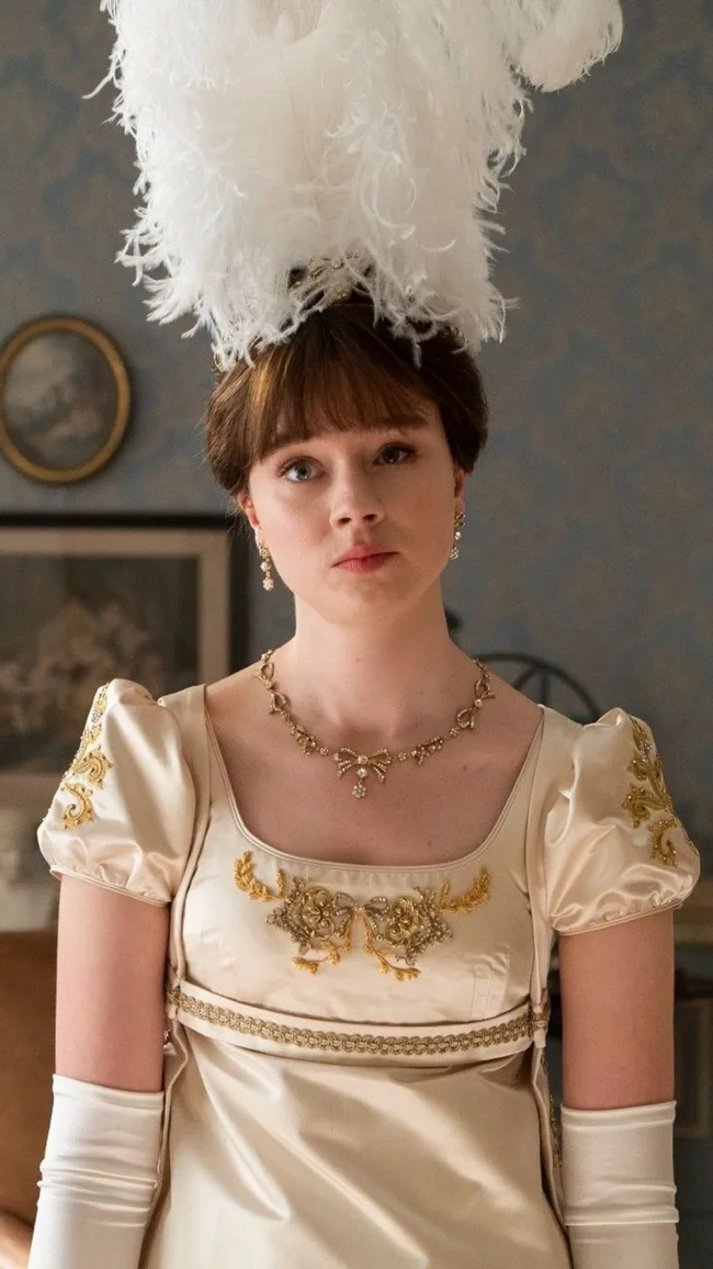 Young woman in ornate cream dress, white gloves, and large feather headdress, in a period setting.