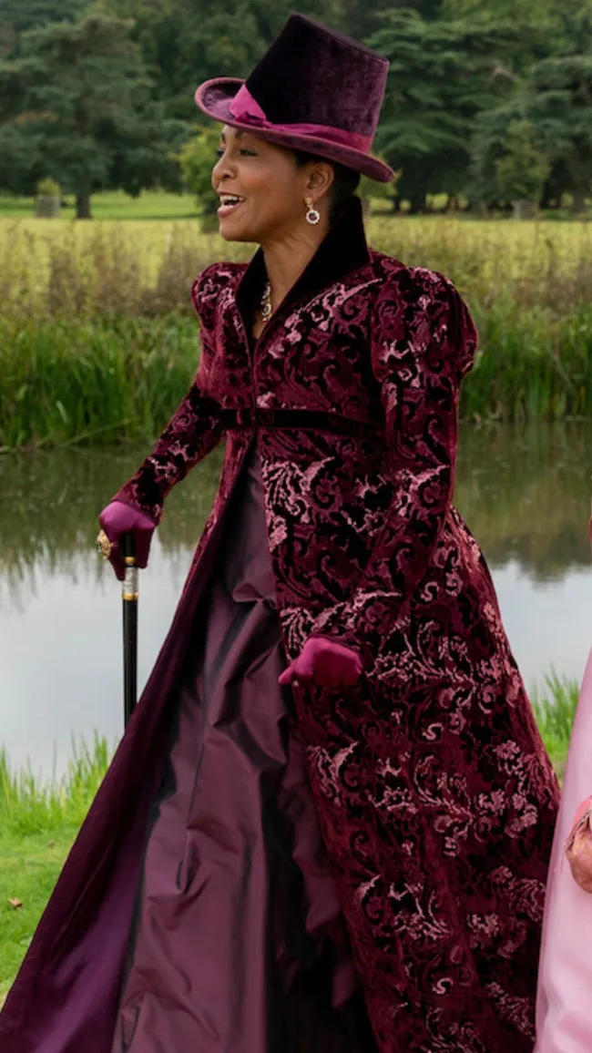A person in a rich, burgundy Victorian outfit with a top hat and cane, standing outdoors.