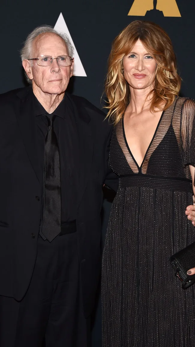 Man in a black suit and woman in a black sheer dress posing together on red carpet event.