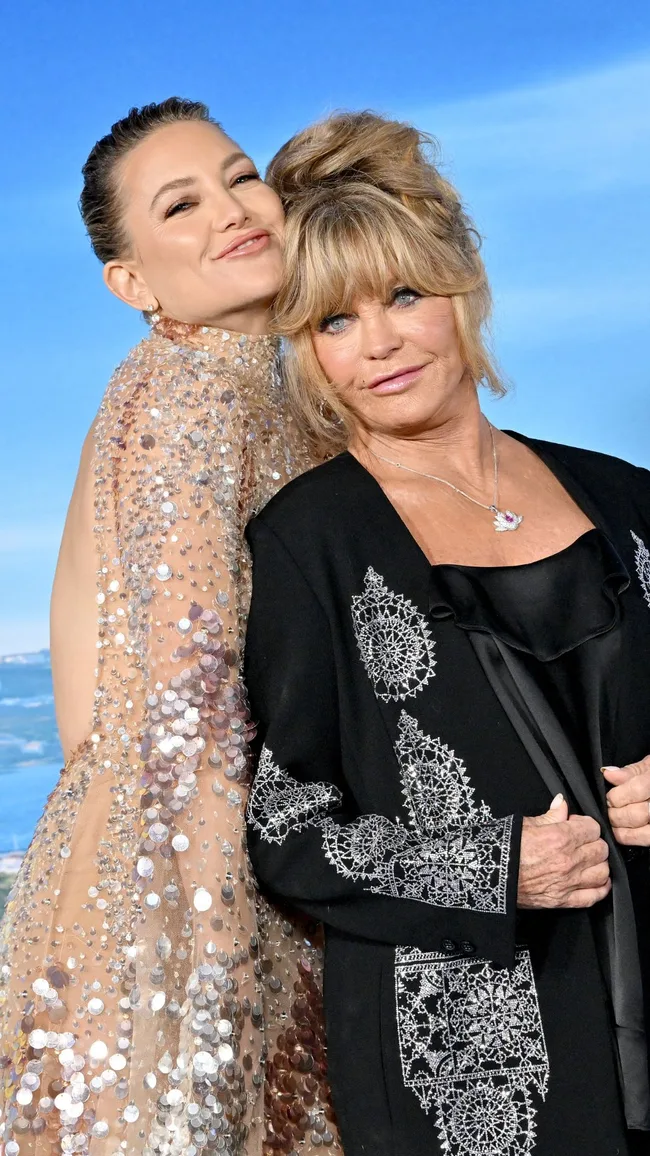 Two women posing together, one in a sparkly dress and the other in a black embroidered jacket, against a blue sky backdrop.