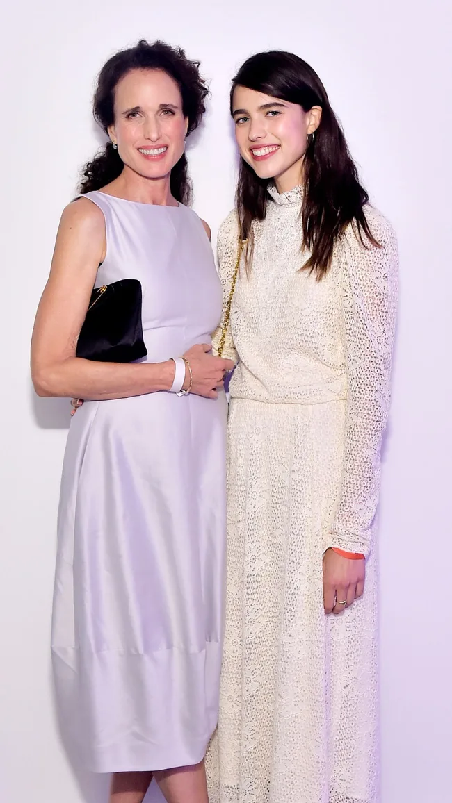 Two women smiling and posing together; one in a sleeveless dress, the other in a long-sleeved lace outfit.