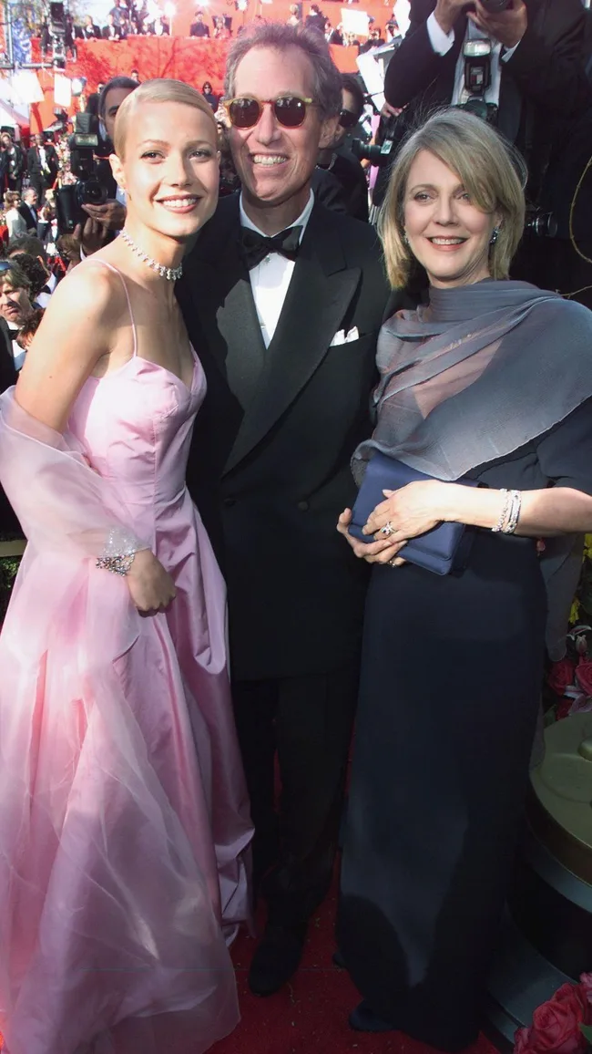 Three people in formal attire smiling on a red carpet, with a crowd and photographers in the background.