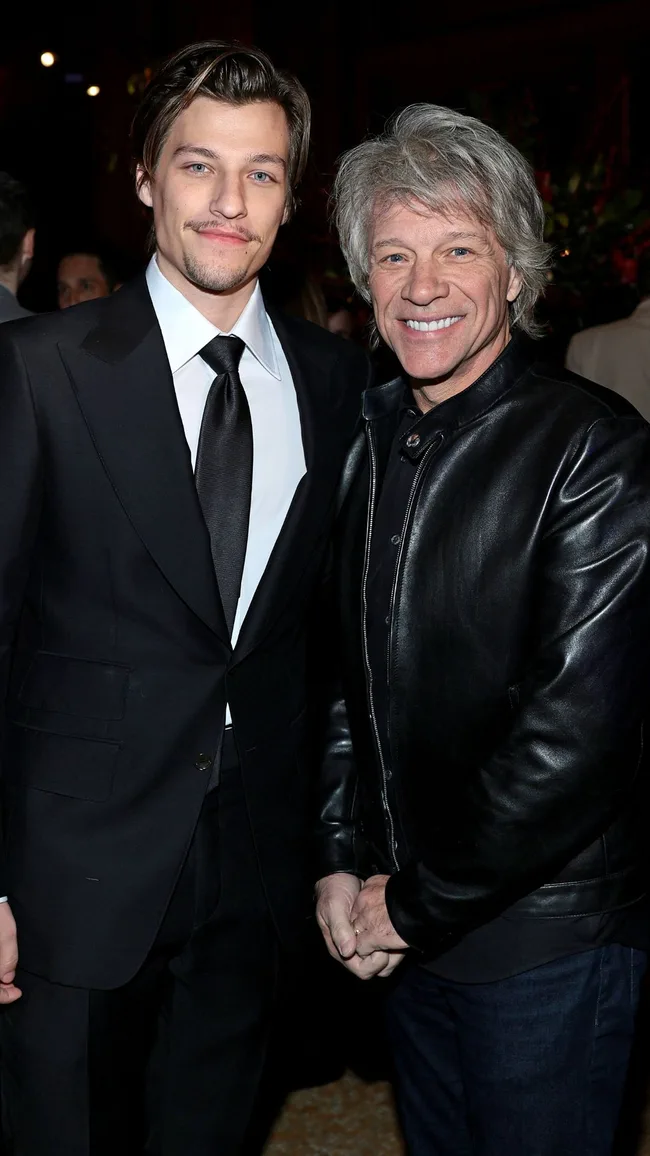 Two men posing together, one in a suit and the other in a leather jacket, both smiling at an indoor event.