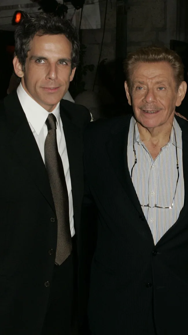 Two men in suits smiling, standing closely together at an event, with dim indoor lighting.
