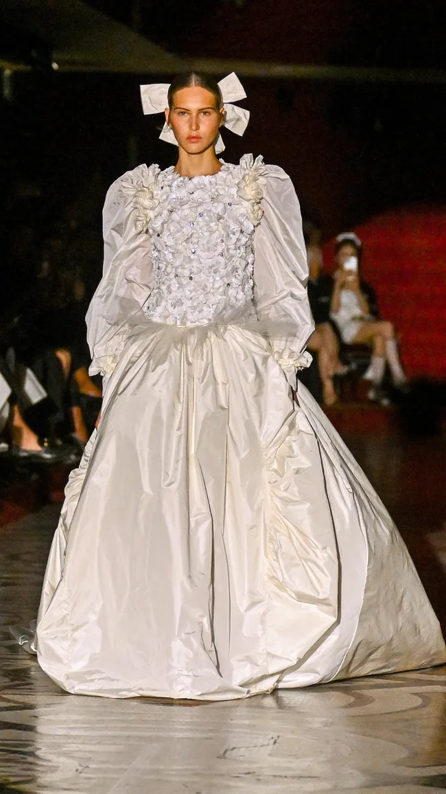 Model in a voluminous white gown with floral embellishments and large hair bows walks a dimly lit runway.
