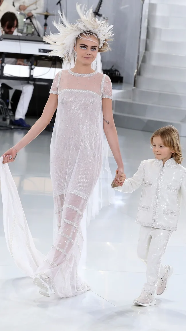 A model in a sheer white dress and feathered hat walks hand in hand with a child in a matching outfit on a runway.