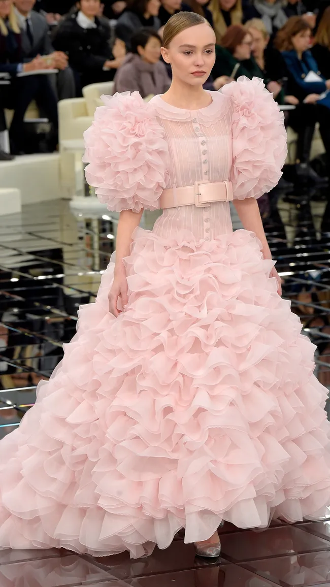 Blonde woman in a ruffled pink gown walks on a reflective runway, surrounded by an audience.