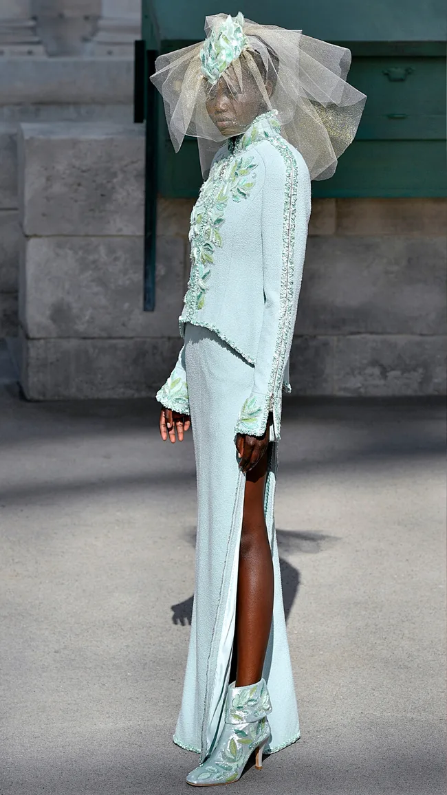 Model in a pale green outfit with floral details and a veil, standing on a concrete surface.