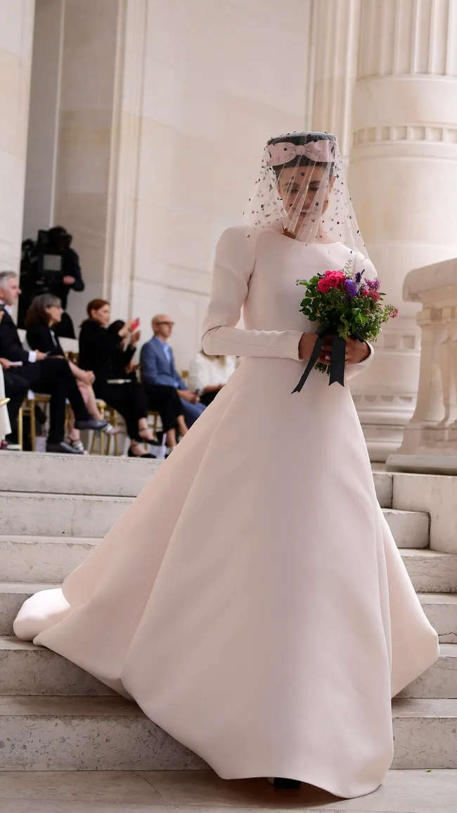 A model in a white bridal gown with a veil walks down stairs holding a colorful bouquet, spectators seated nearby.