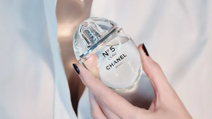 A hand with black nails holding a bottle of Chanel No. 5 L'Eau perfume against a white background.
