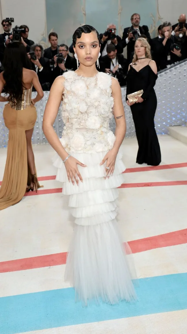 Person in a layered white dress posing on a fashion event red carpet, photographers in background.