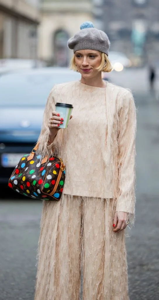 A stylish woman with a short, blonde bob wears a grey beret, peach jump and matching matching skirt. She carried a small louis vuitton bag, which is brown with colourful polka dots, and carries a blue coffee cup.