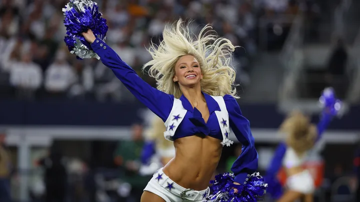 a blonde dallas cowboys cheerleader wears a blue cropped jacket and white shorts, holding a pop pom in the air