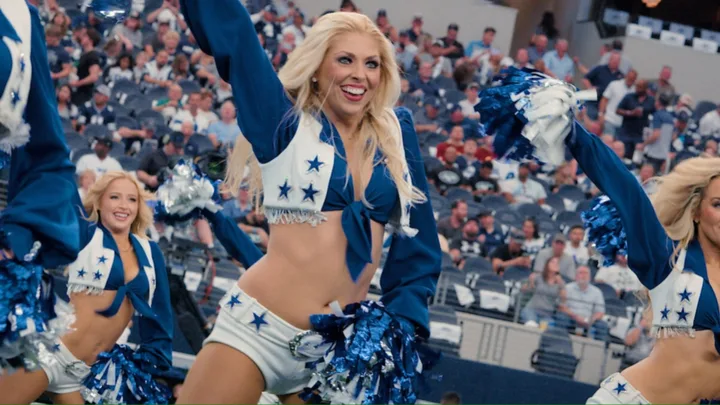 A blonde Dall cowboys cheerleader wears white shorts and a white and blue top, holding pom pons with a hand above her head and one on her hip