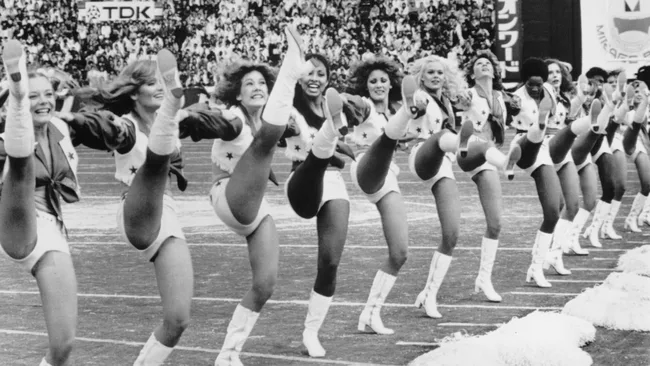 Dallas Cowboys Cheerleaders performing a high-kick routine on a football field, with a crowd in the background.