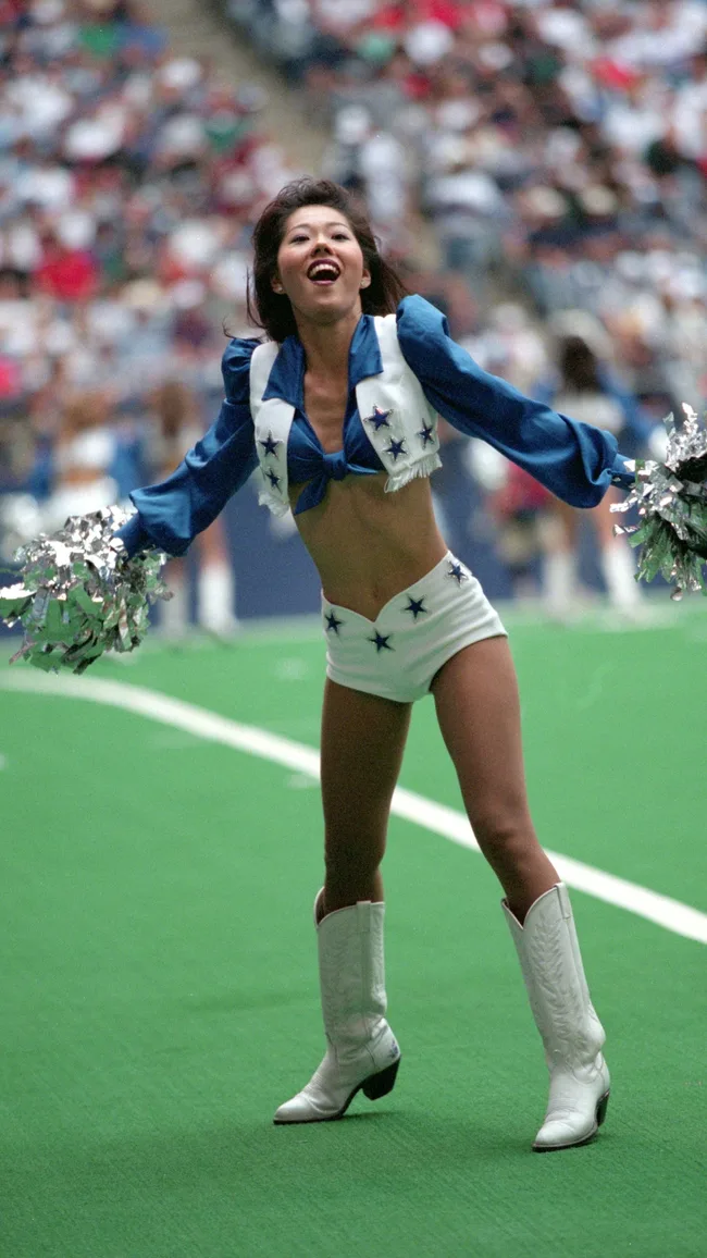 Cheerleader in a blue and white outfit energetically performs at a sports event on a green field.