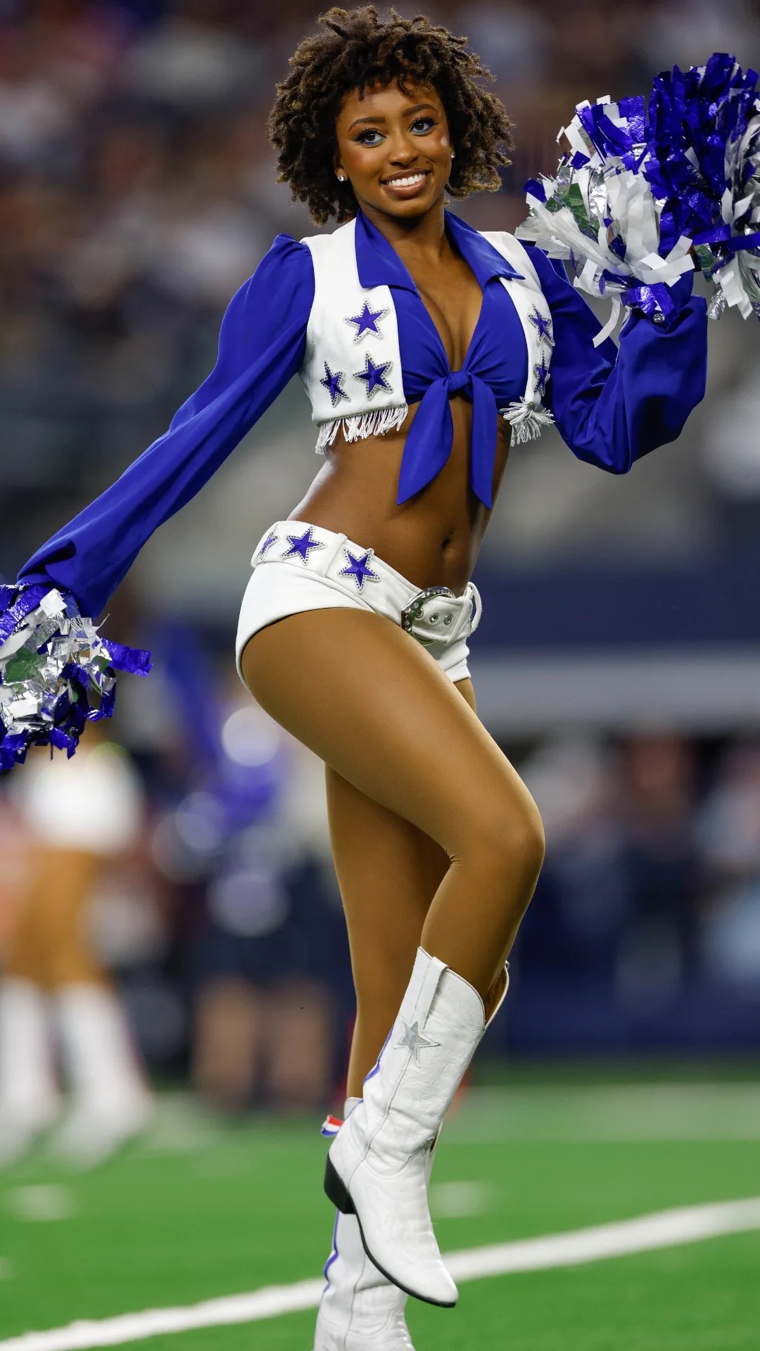 a dallas cowboys cheerleader with a small afro wears a blue cropped short, white vest, white shorts and white boots; she holds silver and blue pom poms