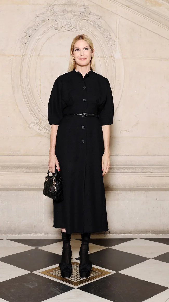 A person in a knee-length black dress with black handbag stands on a checkered floor in front of decorative wall.