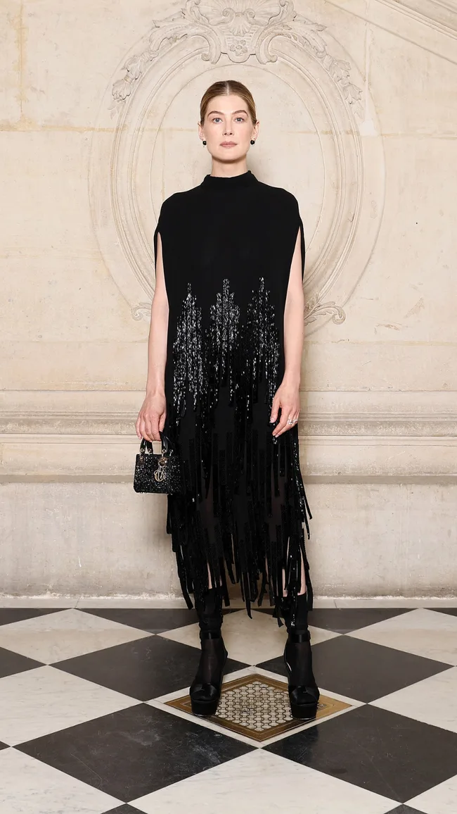 A person in a black sequin dress stands on a black and white tiled floor, holding a small black handbag.