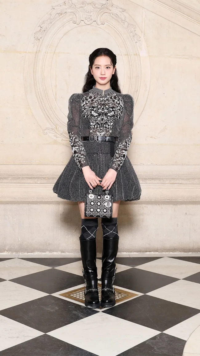 A person in a black and silver dress with boots, holding a decorated handbag, standing on a checkered floor.