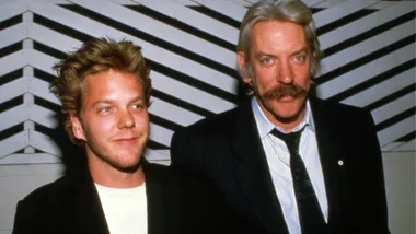 Two people standing, one with blond hair, the other with a mustache, in front of a geometric patterned wall.