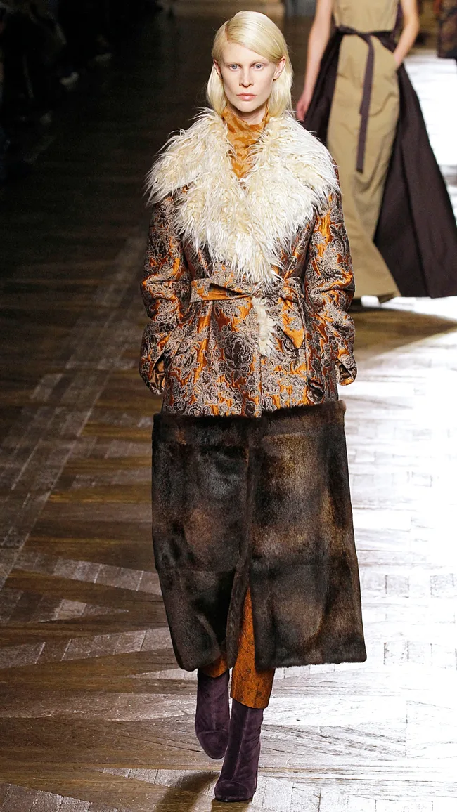 Model in ornate coat with fur detail and long skirt on runway.