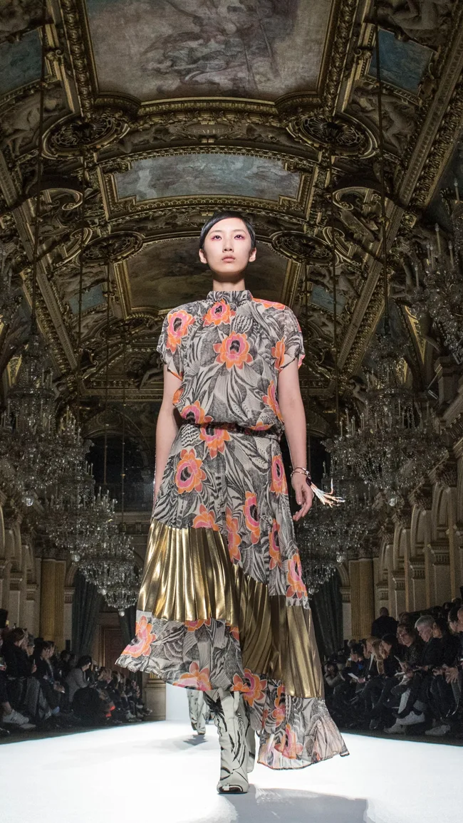 Model in a floral and gold dress walks a runway in an opulent hall with chandeliers and ornate ceiling.