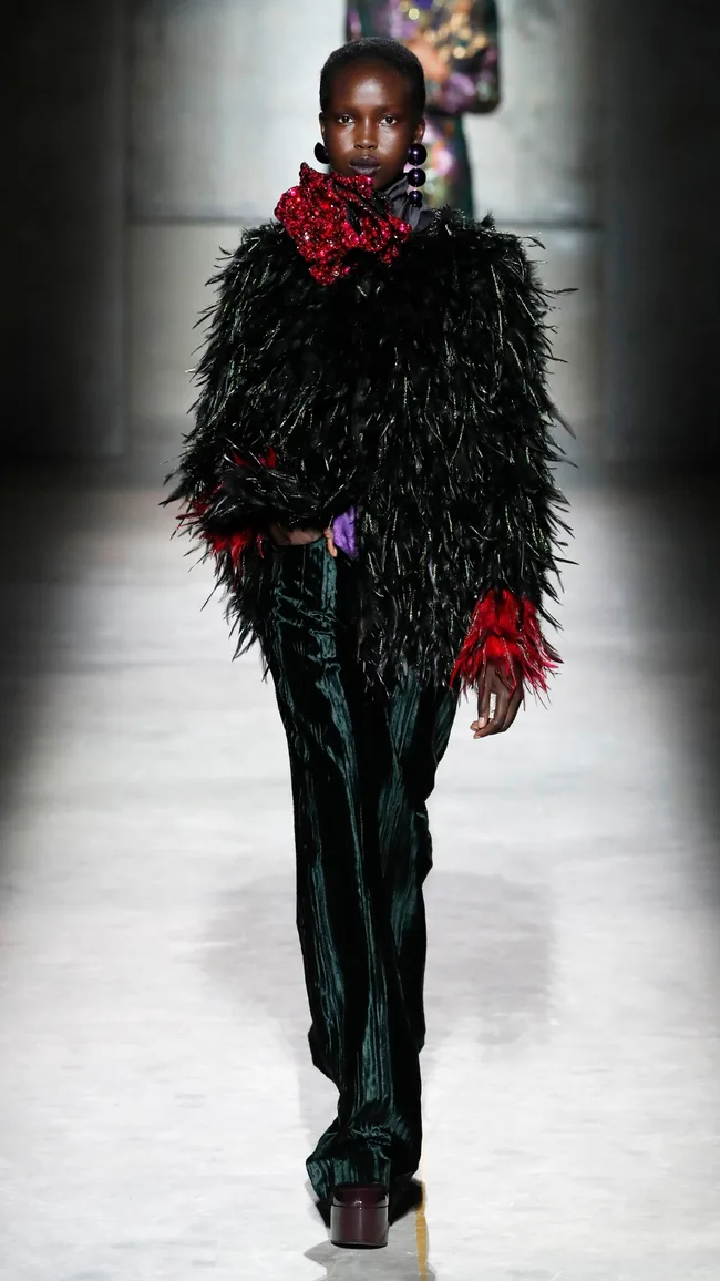 Model on runway wearing a black feathered top with red accents, green velvet pants, and bold earrings.